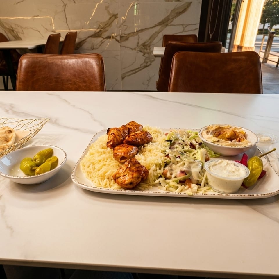 Chicken shish tawook plate with rice, salad, hummus, pita, and pickles on a marble table.