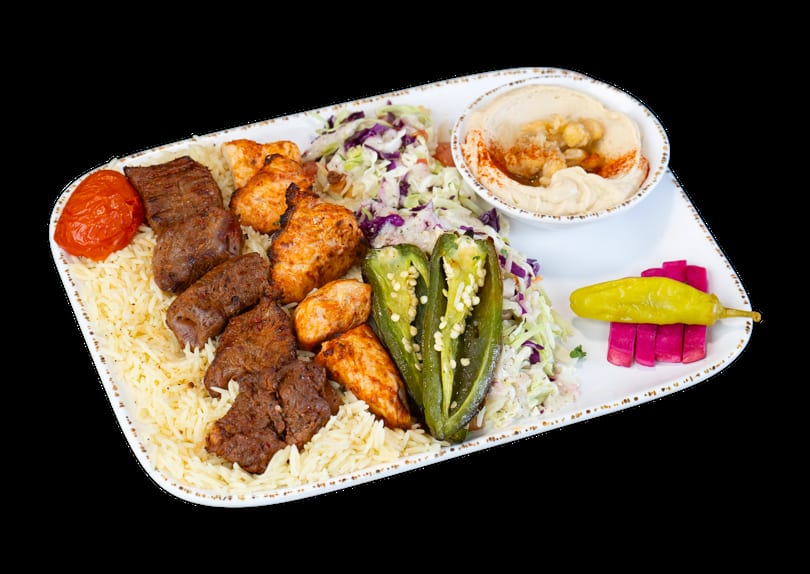A platter with rice, beef and chicken kebabs, coleslaw, hummus, grilled peppers, and pickled vegetables.