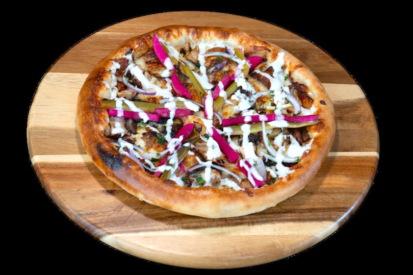 Shawarma pizza with chicken, red onions, pickles, and white sauce on a wooden board.