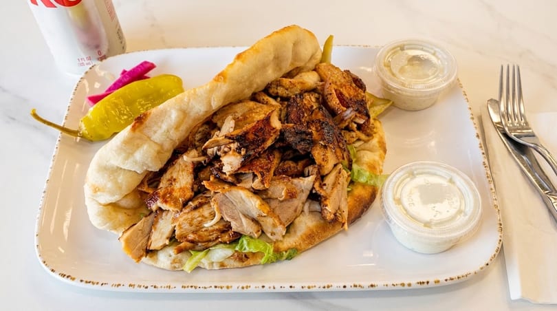 Chicken shawarma on pita bread with lettuce, pickles, and two sauce cups on a white plate.